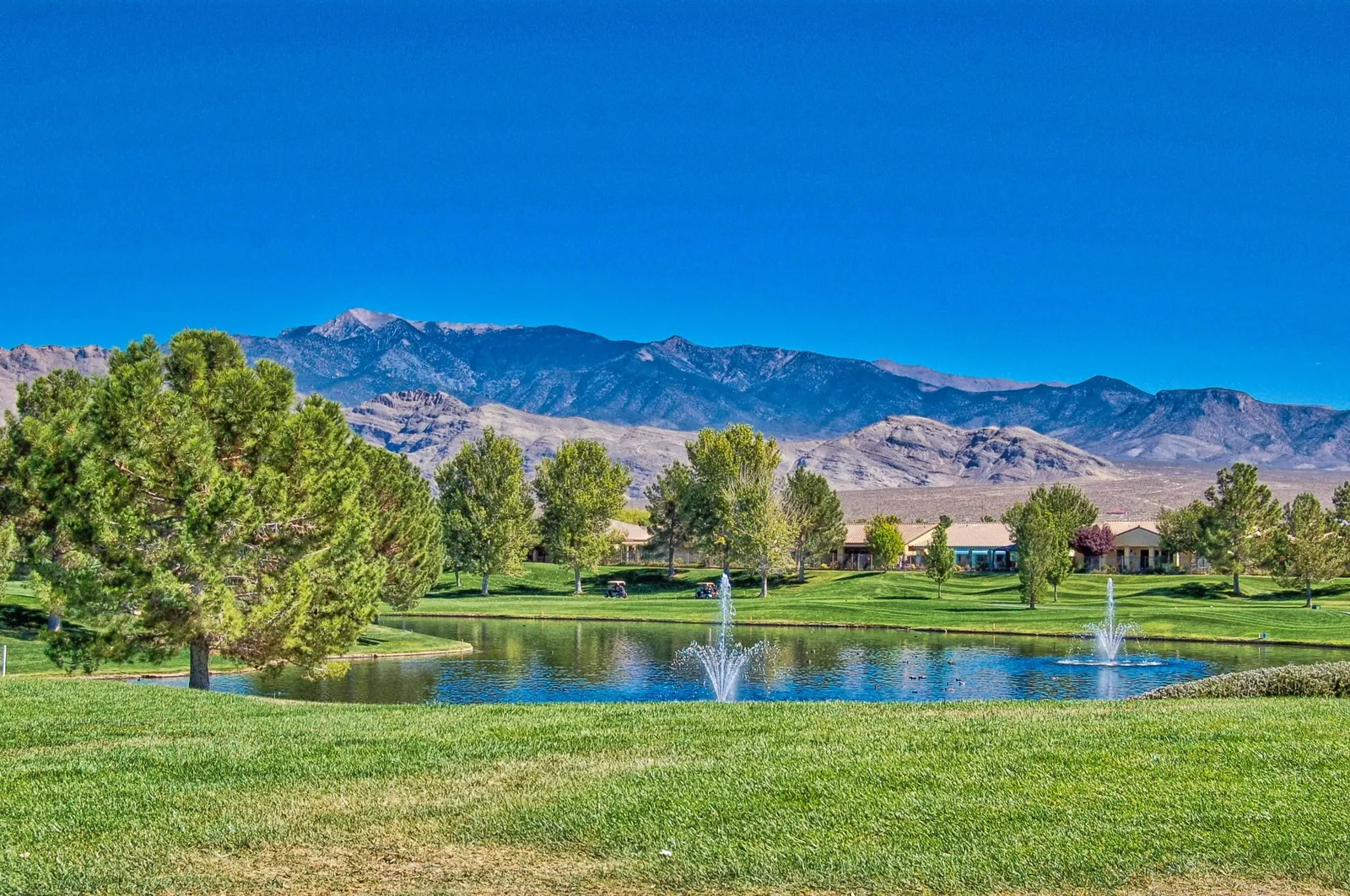 A pond with fountains on a Pahrump golf course, with the Spring Mountains behind under a clear blue sky