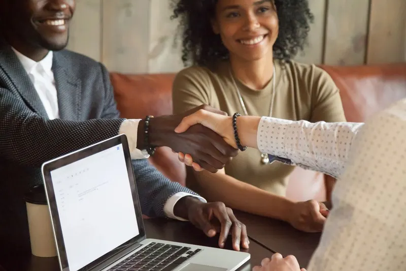 Two people shaking hands across a desk with a laptop